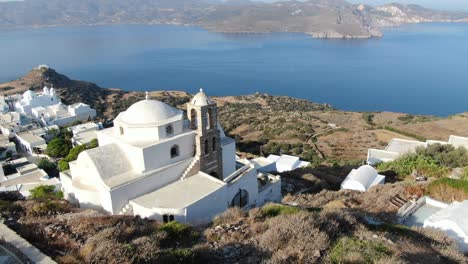 Drone-view-in-Greece-flying-over-a-white-church-on-a-hill-with-a-greek-white-house-town-facing-blue-sea-on-a-mountain