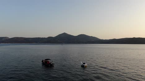 Vista-De-Drones-En-Grecia-Volando-Sobre-El-Mar-Azul-Con-Barcos-Montañas-En-El-Horizonte-En-Milos-Al-Atardecer