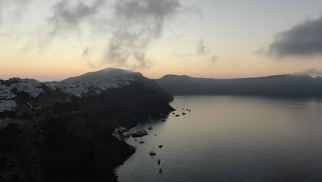 Drone-view-in-Greece-flying-over-Santorini-with-Oia-town-white-houses-on-a-cliff-next-to-the-mediterranean-sea-at-sunrise