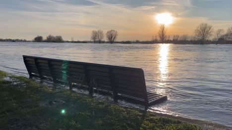 Waves-coming-in-on-the-city-boulevard-riverbed-of-a-flooded-river-IJssel-reaching-a-bench-during-rising-and-high-water-levels-at-sunset-with-vast-floodplain-landscape-trees-under-water-on-the-horizon
