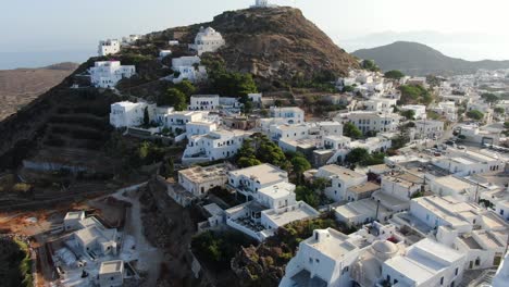 Drone-view-in-Greece-flying-in-front-of-greek-town-with-white-houses-on-a-brown-hill-with-a-church-at-the-top-and-sea-on-the-horizon-on-a-sunny-day