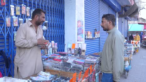 Toma-Cinematográfica-De-Un-Comerciante-Que-Vende-Fundas-Para-Teléfonos-Móviles-A-Su-Cliente-Con-Chaqueta-Durante-Una-Mañana-De-Invierno-En-La-Calle-Saddar-Bazar-De-Karachi.