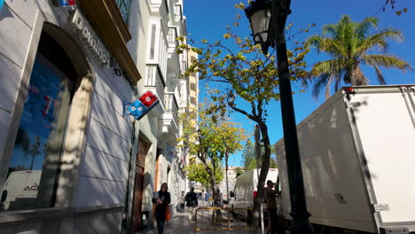 Eine-Malerische-Straße-In-Cádiz-Mit-Traditioneller-Architektur-Und-Einem-Hellen-Werbebanner-Vor-Dem-Hintergrund-Eines-Blauen-Himmels-Und-Baumwipfeln