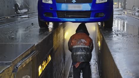 Mechanic-in-raincoat-cleaning-a-car-from-below-with-a-pressurized-water-hose-in-a-car-cleaning-warehouse