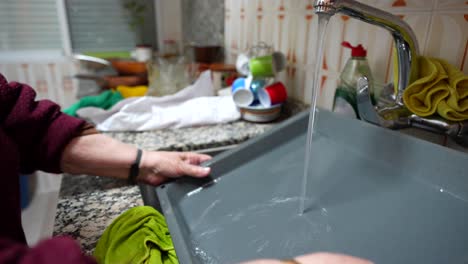 Old-woman-rinsing-a-grey-baking-tray-in-the-kitchen-with-tap-water