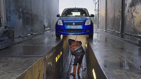 Person-cleaning-the-undercarriage-of-a-blue-vehicle-using-a-high-pressure-water-hose-while-adorned-in-a-protective-raincoat-at-an-automotive-cleaning-facility