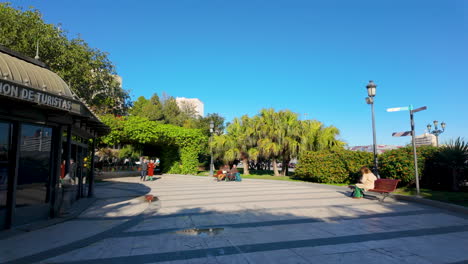 A-spacious-public-square-in-Cádiz-with-a-clear-blue-sky,-featuring-walking-paths,-benches,-and-lush-greenery-surrounding-an-information-center