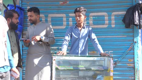 Front-shot-of-a-teenager-boy-working-as-a-labour-near-two-men-talking-to-each-other-during-sunny-afternoon-at-Saddar-Bazar-Street-of-Karachi,-Pakistan