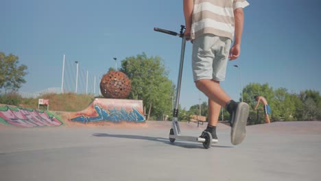 Man-walk-at-public-skatepark-concrete-ground-and-hold-stunt-scooter-beside
