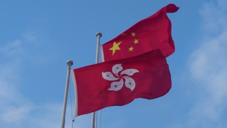 Looking-Up-At-Chinese-And-Hong-Kong-National-Flags-Fluttering-In-Wind-Against-Blue-Sky