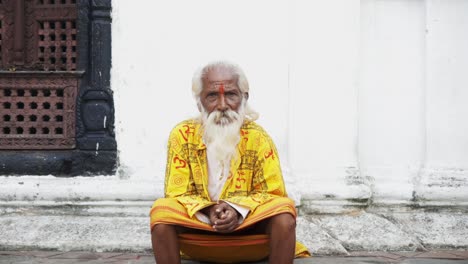 Portrait-shot-of-old-Nepalese-male-monk-sitting-on-stairs-in-front-of-temple-in-Nepal,-close-up