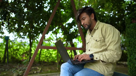 Nepali-Bungo-male-sits-in-the-park-of-Nepal,-writes-content-on-Laptop,-studies-in-green-environment-drone-shot,-advertising-4K