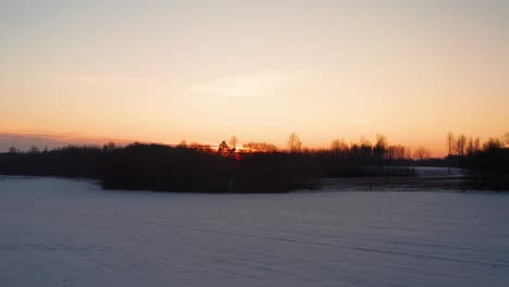 Fliegen-über-Verschneitem-Landwirtschaftlichem-Feld-In-Richtung-Sonnenuntergangswaldsilhouette