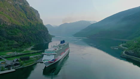 Cruise-ship-sailing-through-a-narrow-fjord-with-towering-mountains,-calm-waters-reflecting-the-serene-landscape,-aerial-view