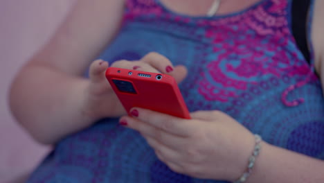Close-up-of-a-fat-woman-scrolling-and-playing-with-her-red-Smartphone-while-sitting,-wearing-a-colourful-blue-pink-dress-and-red-nail-polish-in-slow-motion