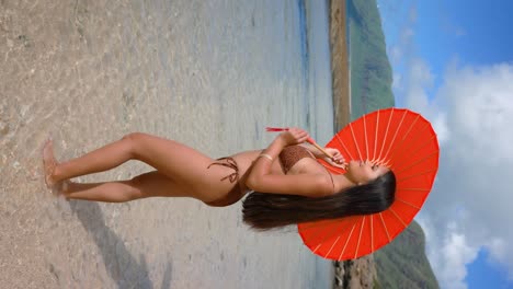Under-the-shade-of-a-red-umbrella,-a-bikini-model-with-long-black-hair-brings-a-chic-lifestyle-concept-to-the-tropical-beach