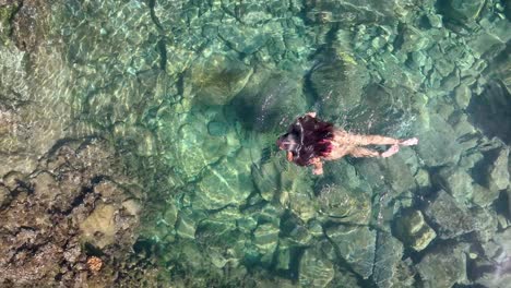From-above,-a-female-model-in-a-swimsuit-unwinds-in-a-small-pool-by-volcanic-rocks-near-the-ocean