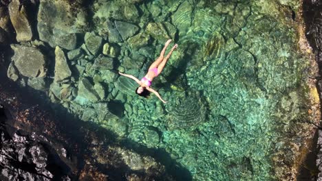 A-swimsuit-clad-model-relaxes-in-a-small-ocean-side-pool-surrounded-by-volcanic-rocks-in-an-aerial-view