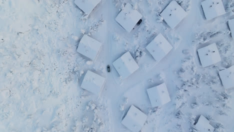 Aerial-birds-eye-shot-following-car-driving-between-snowy-cabins-of-Lapland