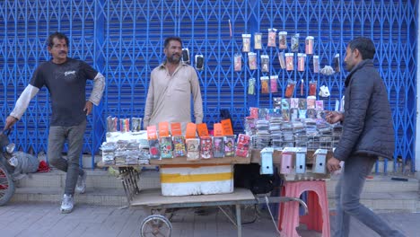 Vista-Frontal-De-Un-Comerciante-Que-Trata-Con-El-Cliente-En-Su-Tienda-De-Carretera-Durante-Una-Mañana-De-Invierno-En-La-Calle-Saddar-Bazar-De-Karachi,-Pakistán.