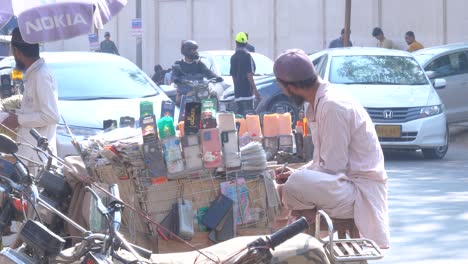 Vista-Lateral-De-Un-Hombre-Revisando-Los-Registros-De-La-Venta-De-Sus-Lentes-Templados-Móviles-En-Su-Tienda-Al-Borde-De-La-Carretera-Por-La-Tarde-En-La-Calle-Saddar-Bazar-En-Karachi,-Pakistán.