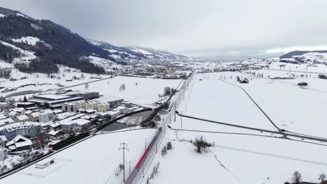 Aerial-view-showing-riding-train-on-Railroad-in-snow-Winter-landscape-and-driving-cars-underpass---Reichenburg,-Switzerland