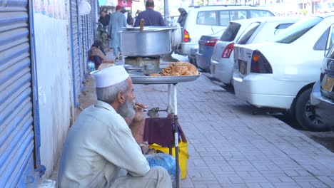 Slow-motion-shot-of-a-hardworking-old-shopkeeper-smoking-beside-his-stall-at-a-roadside-of-Saddar-Bazar-Street-of-Karachi,-Pakistan
