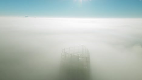 Drone-tilts-up,-unveiling-lone-skyscraper-piercing-through-thick-clouds-at-sunrise—a-cinematic-marvel-in-the-empty-cityscape