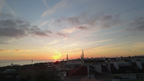 Luftaufnahme-Eines-Sonnenuntergangs-In-Der-Bucht-Von-Lissabon,-Eingebettet-Zwischen-Häusern-In-Der-Nähe-Des-Flusses-Tejo,-Der-Den-Himmel-Und-Das-Wasser-Mit-Warmen-Farbtönen-Inmitten-Der-Stadtlandschaft-Bemalt