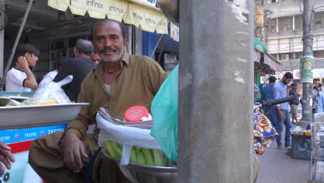 Slow-motion-parallax-shot-of-a-happy-man-selling-chaats-at-roadside-on-a-motor-cycles-during-morning-at-Saddar-Bazar-Street-of-Karachi,-Pakistan