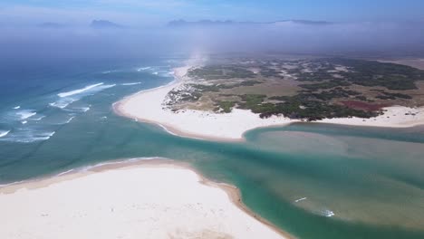 Dramatic-drone-scene-of-a-river-mouth-opening-up-to-the-ocean-with-sand-dunes-and-mountains-in-the-distance