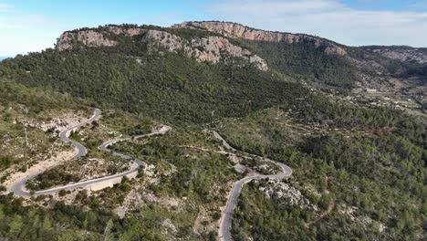 Aerial-approaching-shot-of-mountain-range-in-Esporles-with-Serpentine-Road-on-hill-during-sunny-day,-Mallorca