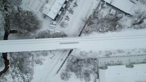 Aerial-top-down-overview-of-train-tracks-crossing-above-road-passing-homes-in-the-winter-snow