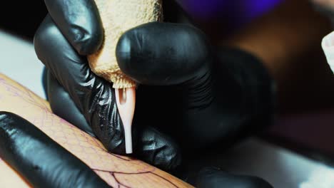 Close-up-slow-motion-tilt-down-shot-of-the-needle-of-a-tattoo-artist-penetrating-the-skin-as-they-follow-the-temp-outline-of-the-desired-image-in-a-dark-studio-room