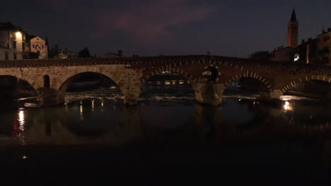 Aerial-Drone-Shot-passing-throught-Ponte-Pietra-Bridge-in-Verona-at-Night-close-to-Adige-River