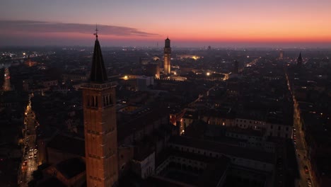 Aerial-Pullin-Drone-Shot,-over-Verona-City-Center-at-Night-with-Sunset-through-Bell-Towers-till-Erbe's-Square