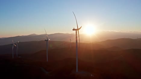 Silhouetted-wind-turbines-on-a-hilly-landscape-with-the-sun-cresting-the-horizon-in-a-serene-aerial-view