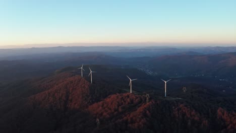 Windkraftanlagen-Krönen-In-Der-Dämmerung-Die-Bewaldeten-Bergrücken-Mit-Fernen-Bergen-In-Einer-Ruhigen-Luftaufnahme