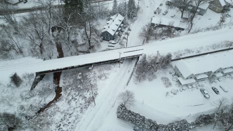 Drone-push-in-above-snow-covered-road-next-to-stream-crossing-below-railroad-tracks
