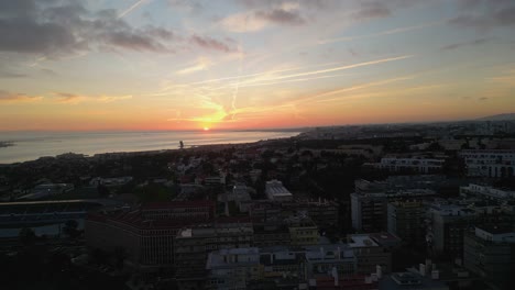 Luftaufnahme-Eines-Sonnenuntergangs-über-Der-Bucht-Von-Lissabon-In-Der-Nähe-Des-Flusses-Tejo,-Der-Warme-Farbtöne-über-Das-Ruhige-Wasser-Und-Die-Stadtlandschaft-Wirft