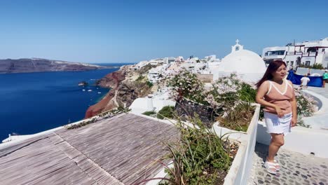 Tourists-Taking-Photos-On-Cliff-|-Oia-Santorini-Greece-Greek-Island-in-Aegean-Sea,-Travel-Tourist-Vacation-Immersive-Moving-Walk-Along-Crowds-Shopping-in-White-Marble-Cliffside-and-City,-Europe,-4K