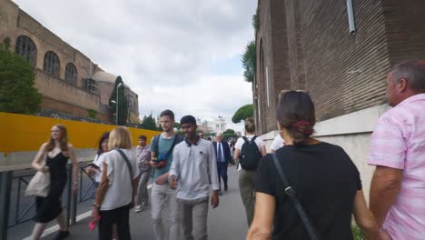 Rome-Immersive-POV:-Moving-In-Busy-Streets-to-Chiesa-Santi-Luca-e-Martina,-Italy,-Europe,-Walking,-Shaky,-4K-|-Elderly-Couple-Holding-Hands-On-Busy-Sidewalk-As-Traffic-Passes