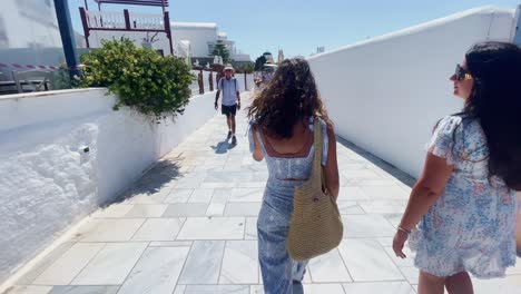 Dos-Mujeres-Caminando-Por-La-Acera-|-Oia-Santorini-Grecia-Isla-Griega-En-El-Mar-Egeo,-Viajes-Turísticos-Vacaciones-Inmersivas-Caminatas-En-Movimiento-Junto-A-Multitudes-De-Compras-En-Acantilados-De-Mármol-Blanco-Y-Ciudad,-Europa,-4k