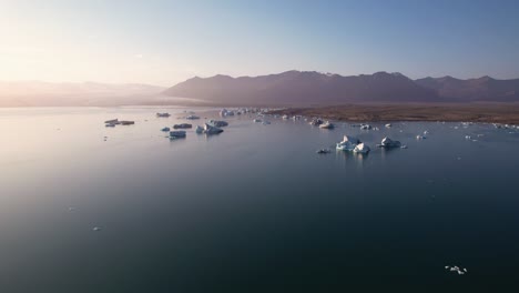Icebergs-Flotando-En-Aguas-Tranquilas-Al-Amanecer-Con-Montañas-Al-Fondo,-Serenos-Y-Majestuosos-Desde-Una-Vista-Aérea-En-Islandia