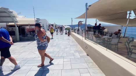 Couple-Walking-Near-Bar-|-Oia-Santorini-Greece-Greek-Island-in-Aegean-Sea,-Travel-Tourist-Vacation-Immersive-Moving-Walk-Along-Crowds-Shopping-in-White-Marble-Cliffside-and-City,-Europe,-4K