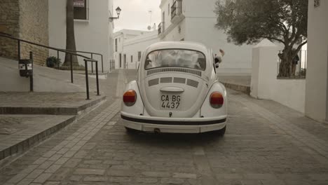 Slow-motion-shot-of-Vintage-Volkswagen-Beetle-driving-people-around-traditional-village,-white-architecture,-cobblestone-road
