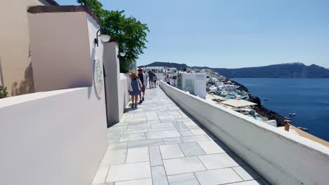 Couple-Walking-Looking-Over-Cliff-|-Oia-Santorini-Greece-Greek-Island-in-Aegean-Sea,-Travel-Tourist-Vacation-Immersive-Moving-Walk-Along-Crowds-Shopping-in-White-Marble-Cliffside-and-City,-Europe,-4K