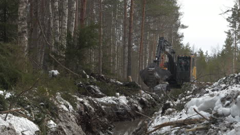 Excavator-clearing-snowy-forest-terrain,-muddy-tracks-among-trees,-mechanical-arm-in-motion,-overcast-day