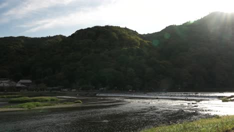 Scenic-Katsura-River-Flowing-Beside-Forested-Hillside-In-Arashiyama,-Kyoto-With-Lens-Flare-In-Shot