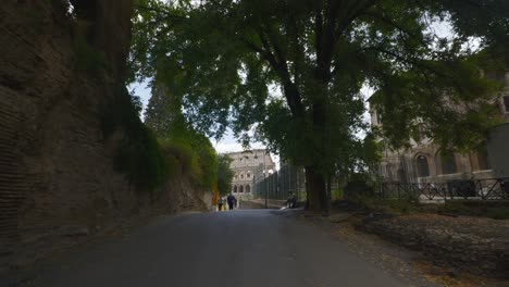 Rome-Immersive-POV:-Moving-In-Busy-Streets-to-Chiesa-Santi-Luca-e-Martina,-Italy,-Europe,-Walking,-Shaky,-4K-|-Tourists-Standing-Near-Street-Vendor-Sitting-On-Path-In-Front-Of-Colosseum